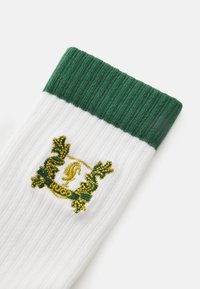 Calcetines blancos acanalados con un puño verde, que tienen un emblema bordado en amarillo y verde etiquetado como "Hugo" en el lateral.