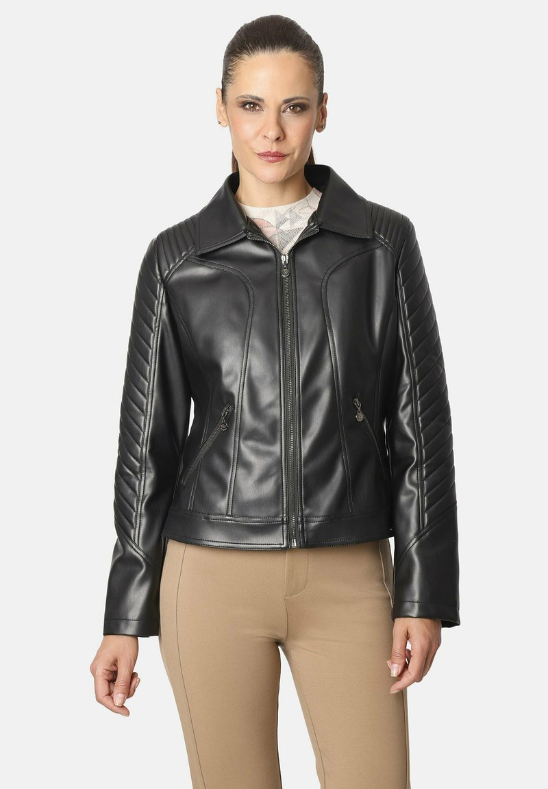 Mujer vistiendo una chaqueta de cuero negra con mangas acolchadas y bolsillos frontales con cremallera, combinada con pantalones beiges, frente a un fondo claro y liso.