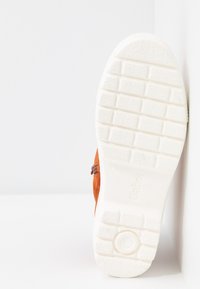Chaussure en daim orange avec une semelle en caoutchouc blanc présentant un motif de grip texturé. Le côté inclut un détail en matériel décoratif.