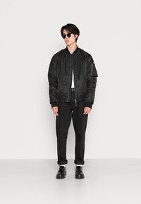 Giacca bomber nera con una texture liscia, polsini e orlo a coste, abbinata a pantaloni e scarpe neri. Modello che indossa occhiali da sole scuri.