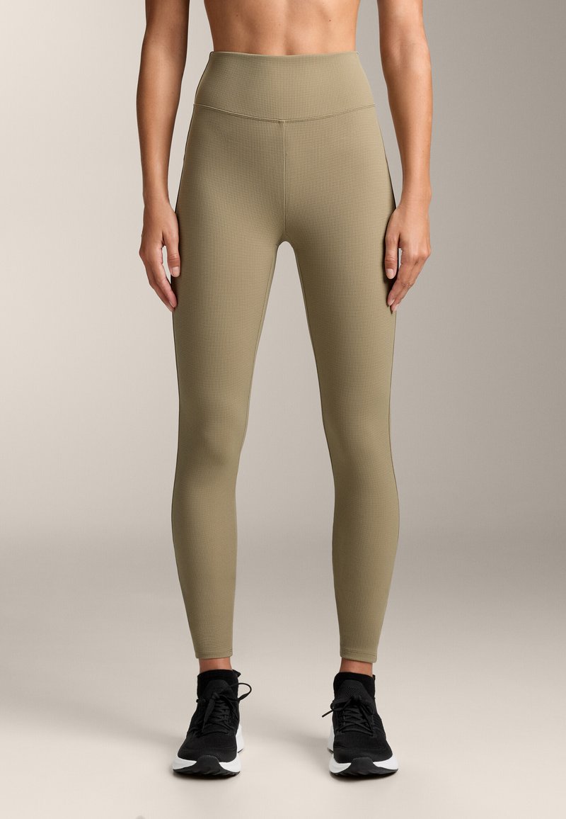 Beige, magas derékú leggings, texturált, bordázott mintával. Rugalmas anyag, szoros illeszkedés, fekete sportcipővel párosítva.
