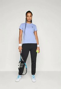 Svetlo purpurna teniška majica s kratkimi rokavi, v kombinaciji z črnimi hlačami, belimi teniškimi čevlji z modrimi detajli, drži teniško lopar in žogico.