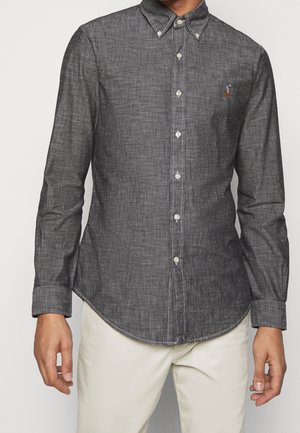 Mężczyzna ubrany w dopasowaną, ciemnoszarą koszulę na guziki z długim rękawem i kołnierzykiem, w połączeniu z jasnobeżowymi spodniami, stoi na jednolitym tle.