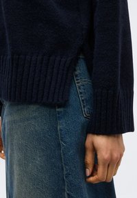 Άτομο που φοράει σκούρο μπλε ριμπ πουλόβερ και μπλε τζιν με εμφανές χέρι που ακουμπάει στο πλάι, κομμένο στη μέση και στο μηρό.