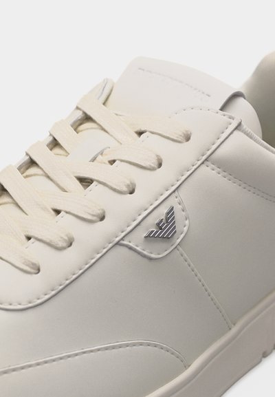 Κοντινή λήψη λευκού δερμάτινου αθλητικού παπουτσιού με κρεμ κορδόνια και μικρό ασημένιο ριγέ λογότυπο στην πλευρική επιφάνεια.