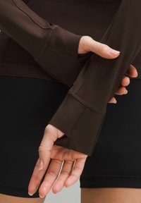 Brown long-sleeve shirt with thumbholes; features flat seams, smooth texture, and fitted design, paired with black shorts for athletic wear.