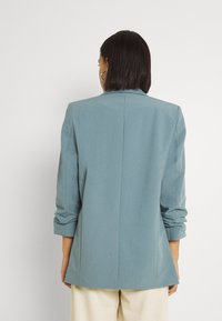 Kobieta z włosami do ramion w ciemnym kolorze nosi jasnoniebieską marynarkę z marszczonymi rękawami oraz kremowe spodnie, odwrócona plecami w stosunku do gładkiego tła.