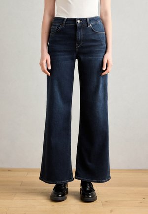 Tmavě modré džíny s vysokým pasem a širokými nohavicemi, nošené s černými mokasíny na dřevěné podlaze, osoba stojící před jednobarevným šedým pozadím.