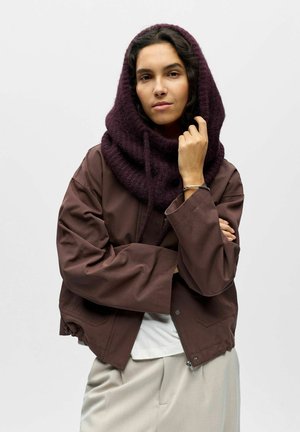 Veste courte marron aux manches larges, superposée à une chemise blanche. Une capuche tricotée de couleur violet foncé est drapée autour du cou, avec une finition texturée.