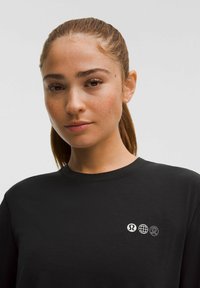 Fiatal nő világos szeplőkkel és hátra tűzött hajjal, fekete pólót visel, amelynek mellkasán három kis fehér logó látható, sima háttér előtt.