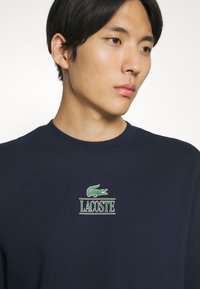 Tmavomodrý sveter s rebrovaným golierom, s výrazným zeleným logom krokodíla Lacoste a textom pod ním. Mäkko štruktúrovaná látka s uvoľneným strihom.