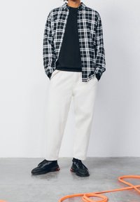 Uomo in piedi con le mani nelle tasche che indossa una maglietta nera, una camicia oversize a quadri bianca e nera, pantaloni bianchi, scarpe da ginnastica nere, accanto a un cavo arancione sul pavimento.