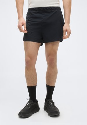 Férfi, fekete sport rövidnadrágban, fekete zokniban és fekete futócipőben, egyszerű fehér háttér előtt.