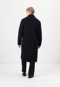 Meripäin villakangastakkia suoran leikkauksen, notkatakin kaulan ja sivutaskujen kanssa. Takana on halkio ja pintakuviointi. Näkyvissä mustat housut ja kengät.