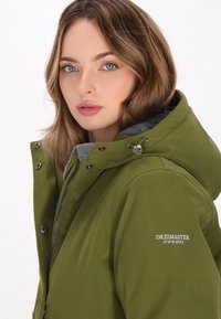 Giacca con cappuccio verde oliva con zip e chiusura a bottone, dotata di fodera grigia, tasche laterali e dettaglio del logo ricamato sul braccio.