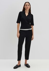 Haut noir à manches courtes avec col, porté par-dessus un sous-pull blanc moulant ; accompagné d'un pantalon noir fuselé et de chaussures à enfiler.