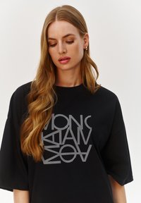 Czarna oversize koszulka z białym graficznym nadrukiem przedstawiającym stylizowany tekst. Materiał wygląda na gładki i lekki, z okrągłym dekoltem.