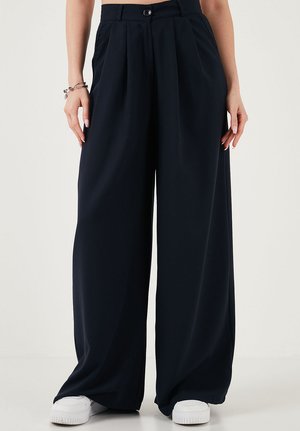 Naine, kes kannab kõrge vöökohaga laia säärtega musti pükse, millel on ees voldid ja küljetaskud, valgete tossude ja vasakul randmel käevõrudega.