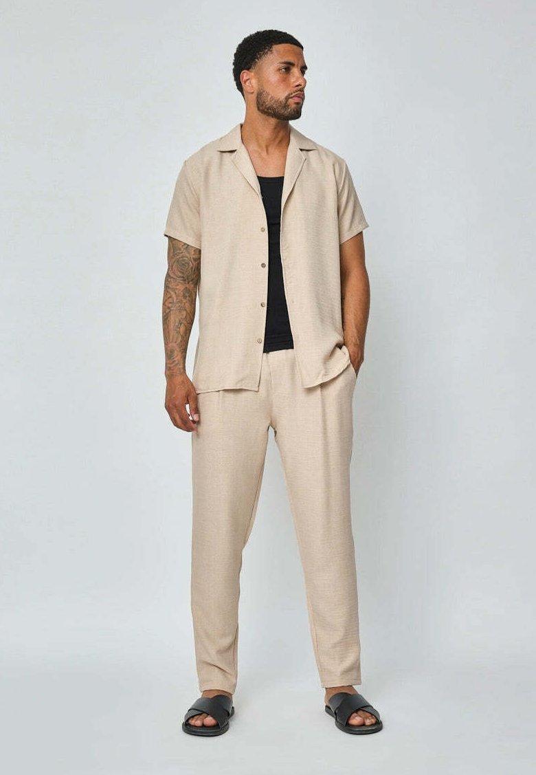 Chemise beige à manches courtes avec col et fermeture à boutons, associée à un pantalon fuselé assorti. Portée avec un débardeur noir et des sandales à enfiler.