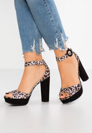 Ben iført slidte blå jeans og åbne højhælede sko med leopardmønstrede remme og tykke sorte blokhæle på hvidt gulv.