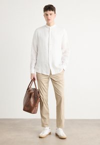 Witte linnen blouse met lange mouwen, beige chinos, witte sneakers met tan kleurige zolen en een bruine leren tas. Staand op een lichtgrijze ondergrond.