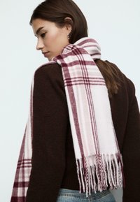 Écharpe à carreaux en tissu doux, avec des rayures bordeaux et rose clair, aux bords frangés. Portée sur un pull marron.
