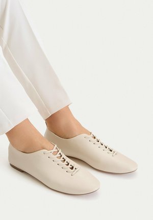 Füße in cremefarbenen minimalistischen Schnürflats, kombiniert mit weißen, knöchellangen Hosen vor einem schlichten Hintergrund.