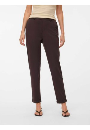 Femme portant un pantalon droit marron foncé avec des revers, un haut beige sans manches et des sandales noires à bouts ouverts, debout devant un fond blanc.