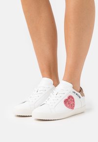 Vita sneakers med rosa glittrande hjärtan och texten "LOVE MOSCHINO" på sidan. Rund tå och lätt sula med bruna detaljer.
