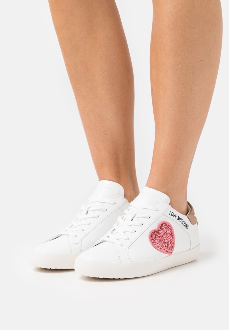 Vita sneakers med rosa glittrande hjärtan och texten "LOVE MOSCHINO" på sidan. Rund tå och lätt sula med bruna detaljer.
