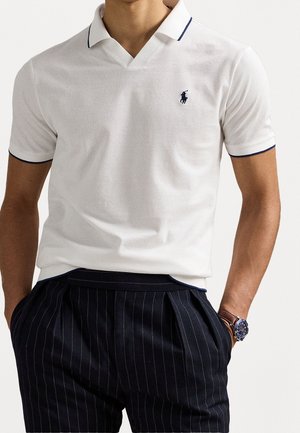 Mann trägt ein weißes Poloshirt mit marineblauem Besatz und Emblem, marineblaue Nadelstreifen-Hosen mit Falten und eine braune Armbanduhr mit blauem Zifferblatt.