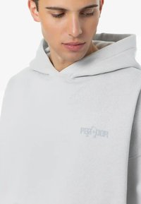 Lichtgrijze hoodie met een zachte textuur, voorzien van een grote capuchon, een kangoeroezak en een ingebrand logo op de borst. Eenvoudige, relaxte pasvorm.