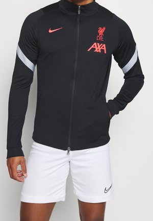 Personne portant une veste de sport noire zippée avec les logos rouges de Liverpool FC et AXA, et un short blanc avec le logo noir Nike, mains près des poches.