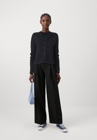 Cardigan azul-marinho com decote em V e fecho de botões, combinado com calças largas pretas com riscas verticais e sapatilhas azuis, segurando uma mala de ganga.