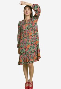 Blumenkleid mit einem dunkelgrünen Hintergrund und lebhaften orangefarbenen und rosa Blumen, langen Ärmeln und einem ausladenden Rüschensaum. Rote Schuhe runden den Look ab.