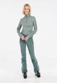 Maglia da sci verde a maniche lunghe con colletto alto e zip, abbinata a pantaloni da sci verdi coordinati. Stivali da sci neri, texture del tessuto liscia.