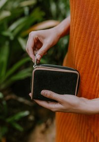 Black suede zippered wallet with a tan leather accent and silver zipper, held in a hand against a backdrop of green foliage.