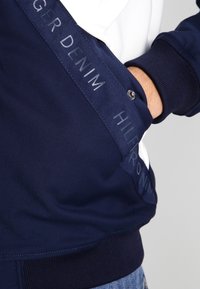 Sudadera azul marino con acentos blancos, que presenta un tejido suave, puños acanalados y un detalle de logo en relieve en un panel lateral.