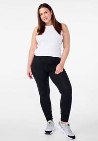 Valge varrukateta top ja mustad retuusid. Retuusidel on sile tekstuur ning modell kannab halli spordijalatsit mustade aktsentidega.