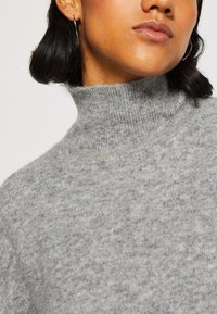 Pull-over turtleneck gris clair en tricot doux avec une texture subtile. Présente un col montant et une coupe décontractée.