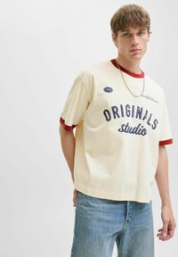 Jauns vīrietis valkā krēmkārtas T-kreklu ar sarkanu apmalīti, ar tekstu "Originals studio" un gaiši zilas džinses, stāv uz viendabīgas baltas fona.