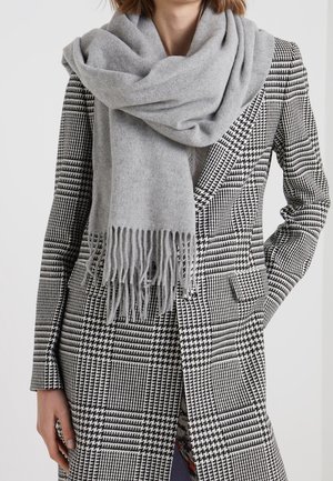 Femme portant un manteau pied-de-poule noir et blanc, les mains dans les poches, avec une grande écharpe gris clair à franges enroulée autour du cou.