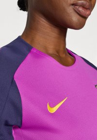 Lilla idrettsdrakt med marineblå detaljer. Har Nike-logo i gult, teksturert stoff og rund halslinning. Nærbilde.