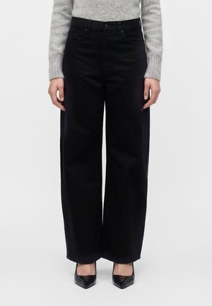 Femme portant un pantalon noir taille haute à jambes larges, associé à un pull gris clair à manches longues et des escarpins noirs pointus.