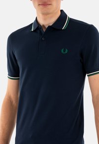 Polo de color marino hecho de tejido texturizado, con un logo de laurel verde y acentos a rayas en el cuello y los puños de las mangas.