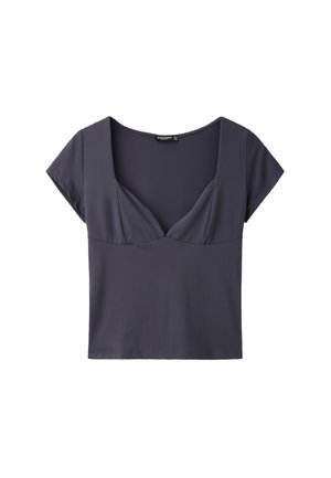 Jeune femme portant des lunettes de soleil foncées, un haut gris cintré à manches courtes, un pantalon large noir avec ceinture et des chaussures noires, debout devant un fond gris uni.