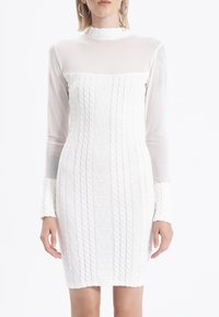 Robe blanche avec manches longues en voile transparent, col montant, et corps en tricot texturé présentant des motifs subtils et des accents côtelés aux poignets.