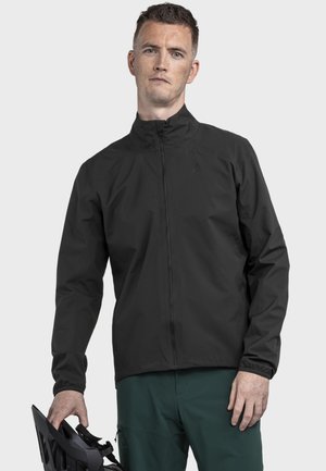 Schwarze, leichte Jacke mit Reißverschluss, hohem Kragen, elastischen Bündchen und glattem Finish, getragen über grüner Hose, mit einem Helm in der Hand.