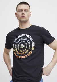 Svart bomull t-skjorte med sirkulært tekstdesign i gradientfarger som sier "Here Comes the Sun" og andre uttrykk, rund hals og korte ermer.
