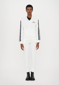 Witte trainingsoutfit met een ritsjack en bijpassende broek. Kenmerkt zich door marineblauwe strepen op de mouwen en een logo-opdruk. Soepele textuur, ontspannen pasvorm. Zwarte laarzen.